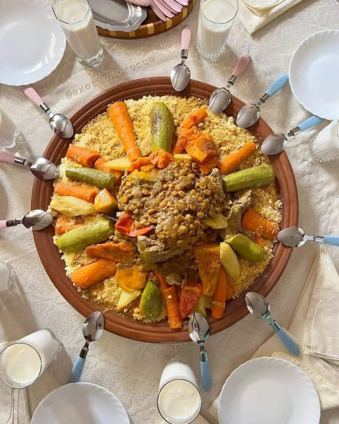 An overhead view of a large couscous platter surrounded by spoons, glasses and individual plates
