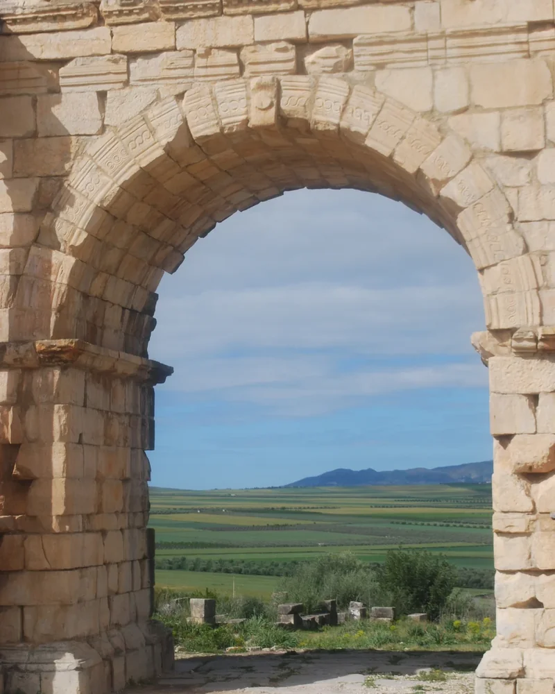 A stone arch at Volubilis frames the surrounding fields.