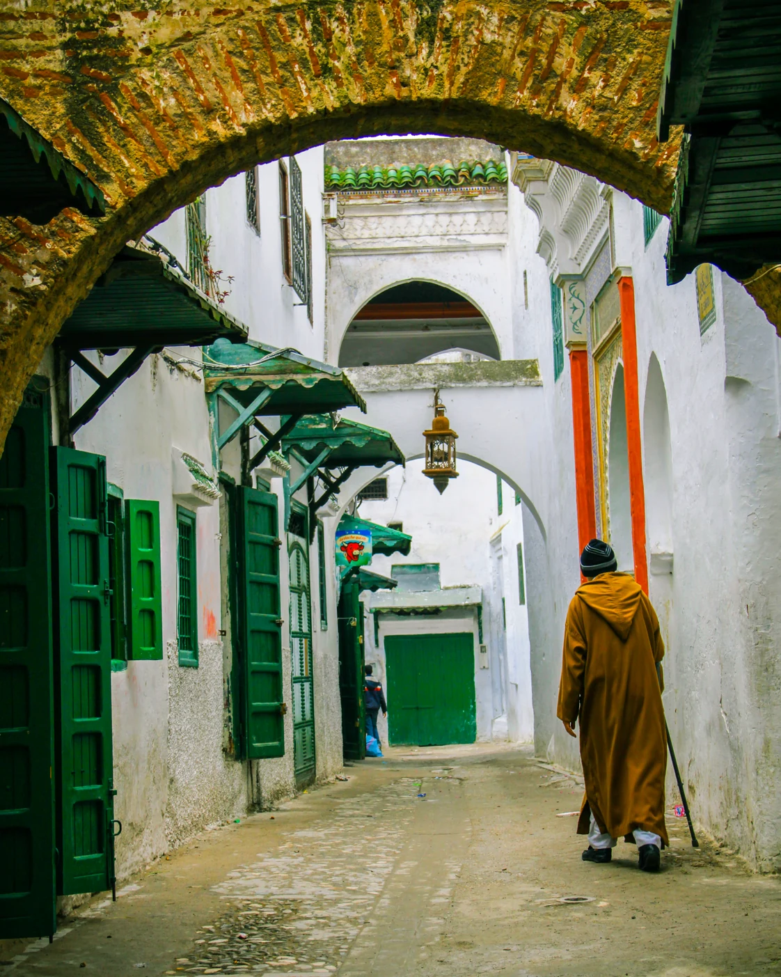 A person in an ochre djellaba walks through a white and green medina lane in Tétouan.