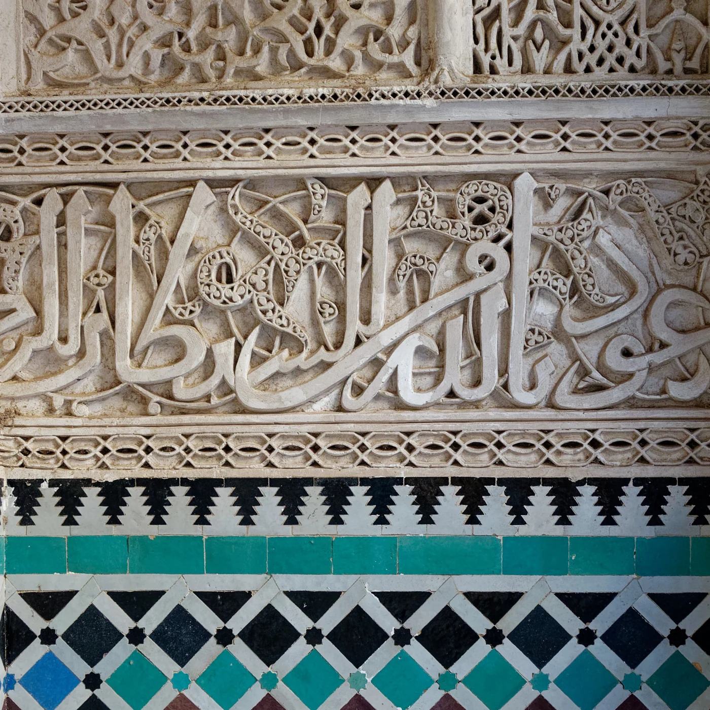 Close architectural detail of turquoise, blue and white zellige beneath carved plaster at Al-Attarine.