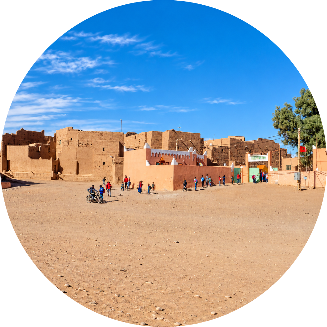 Open village square bordered by earthen buildings, with people crossing the sandy ground