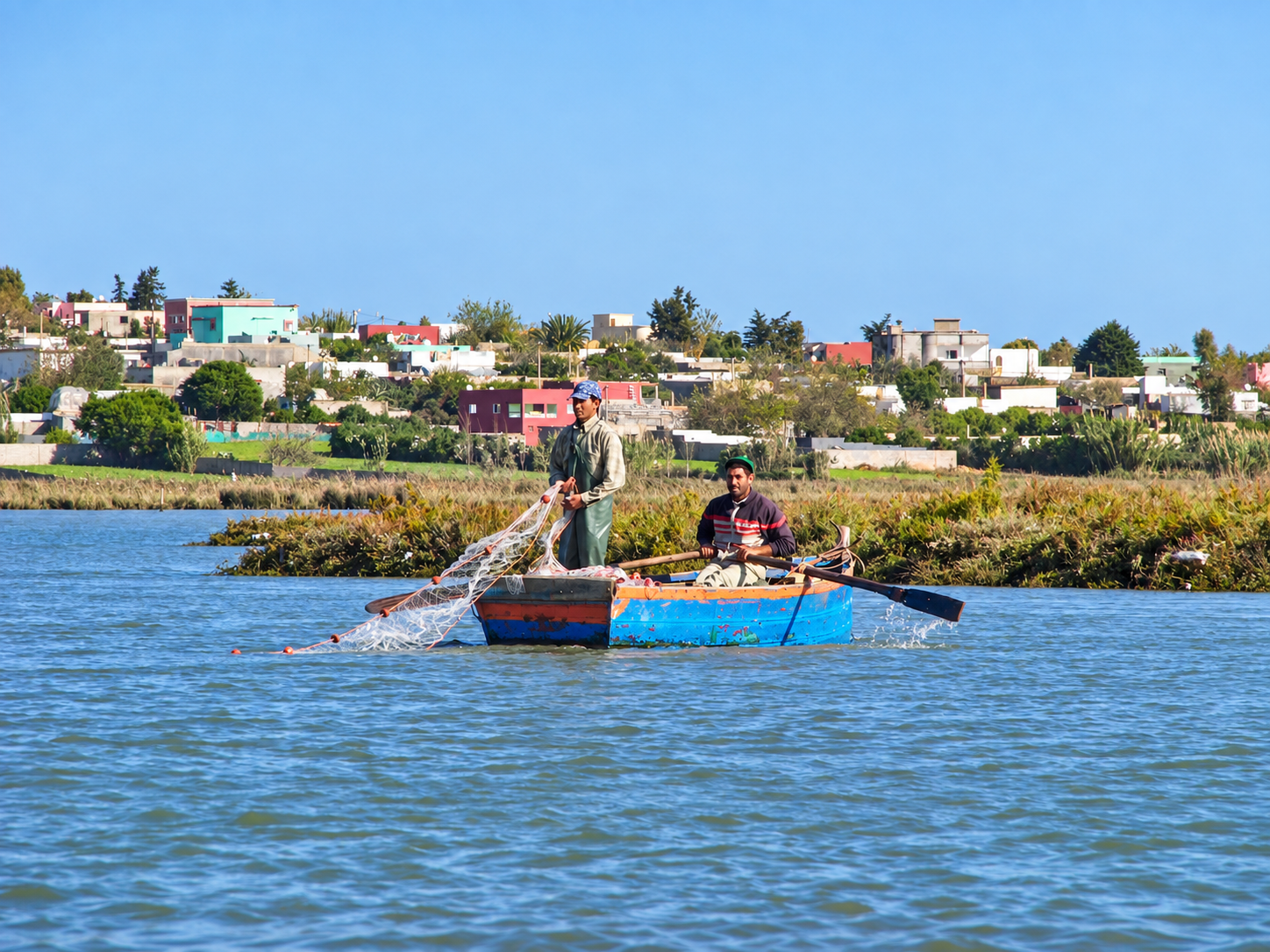 Two fishermen handling a net from a blue boat on the lagoon